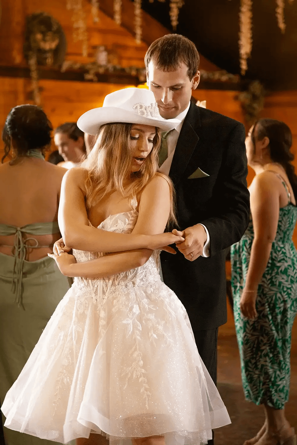 Bride in a cowboy hat adorned with jewels dancing with groom at indoor wedding reception