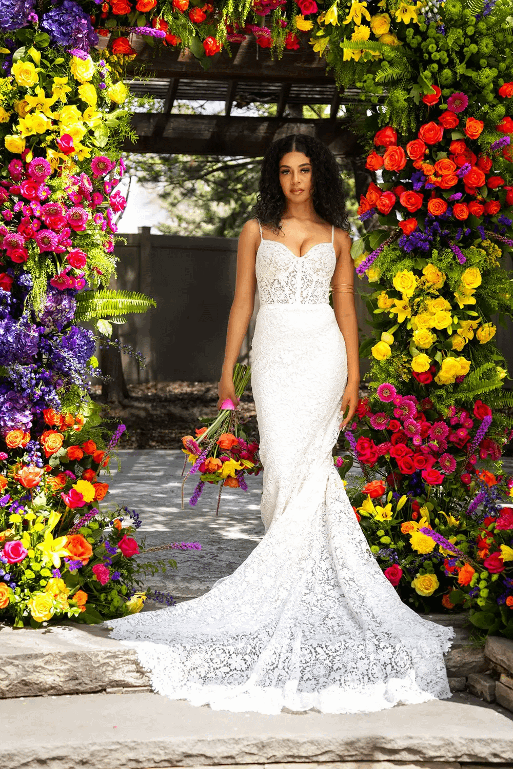 Bridal portrait of a bride in a lace spaghetti-strap gown standing in front of a colorful floral arch